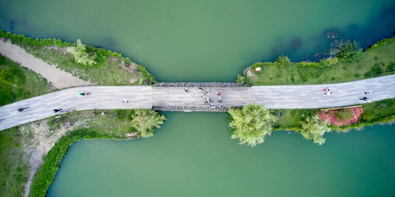 Top down aerial view of the pedestrian bridge in the middle of the pond in the park. People are moving through